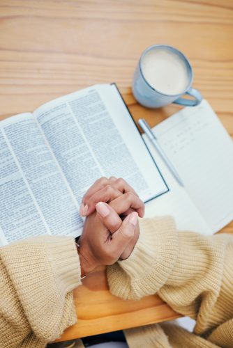 Bible, notes and hands of woman in prayer at desk in home, Christian faith or knowledge of God from