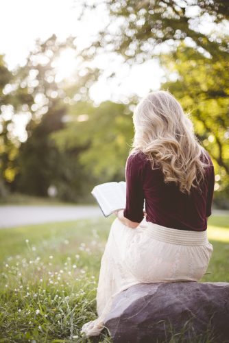 Vertical shot from behind of a female sitting on a rock and reading the bible